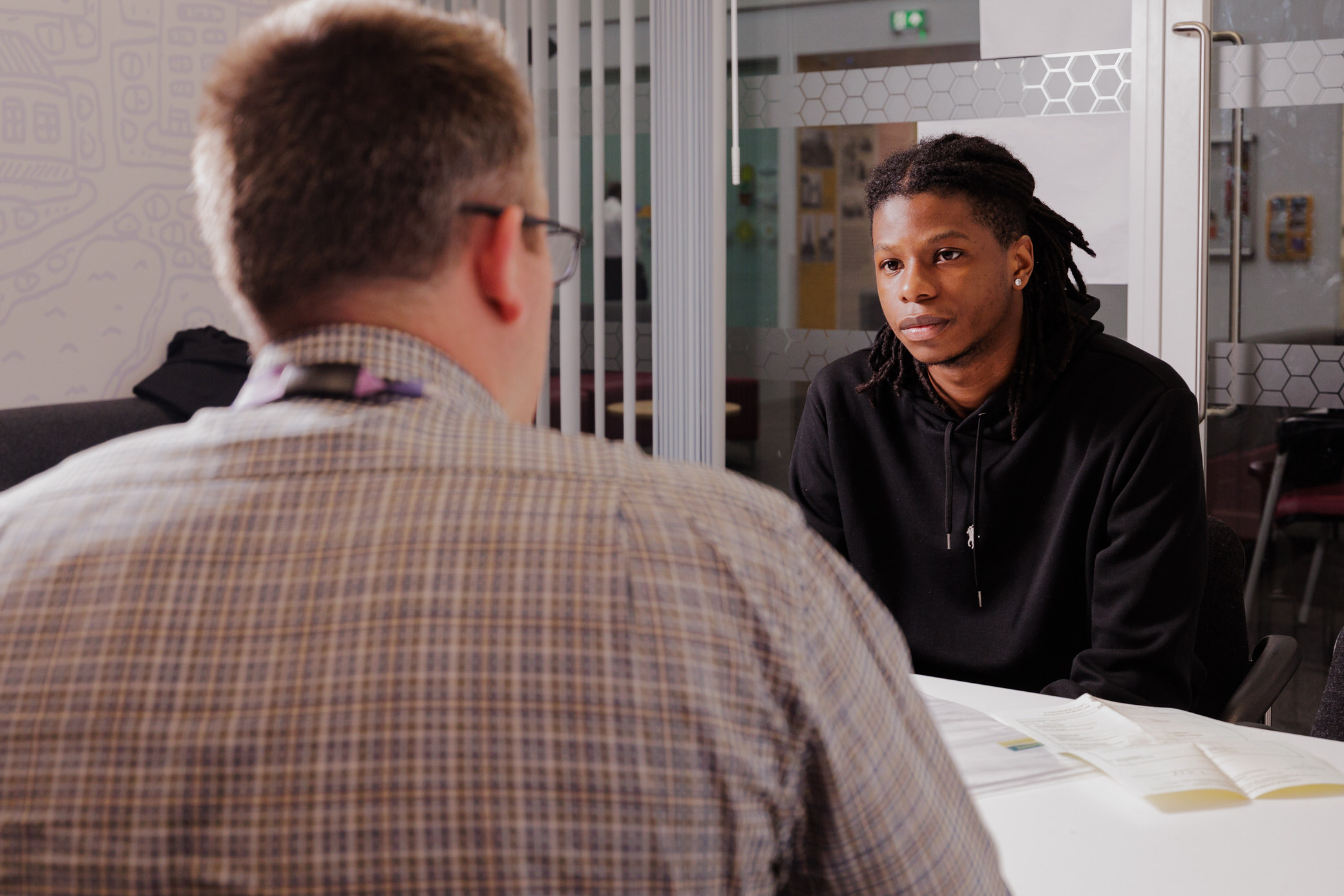 Two people in conversation in an office setting. One is wearing a shirt and the other is wearing a black hoodie. 