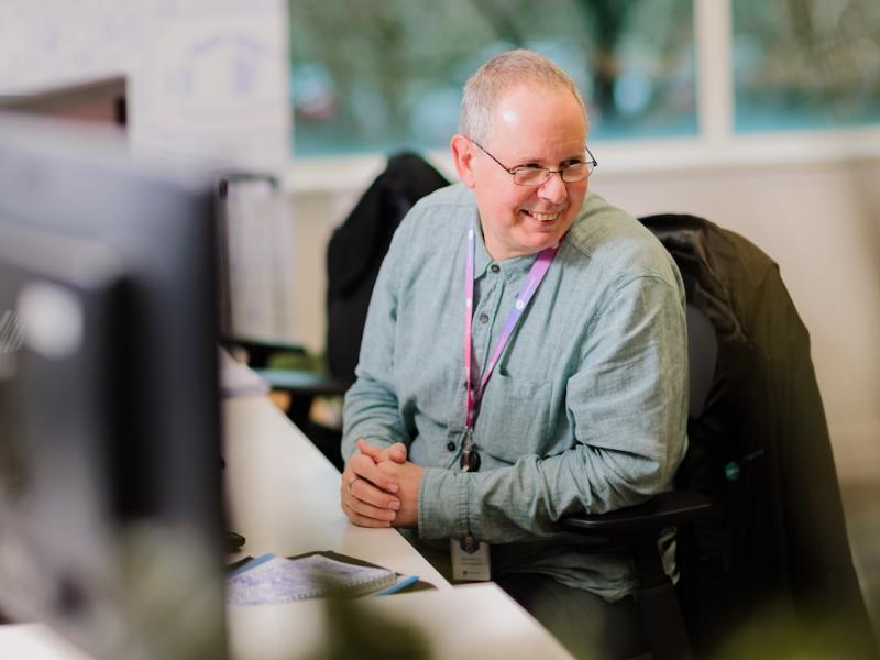 A colleague of One Manchester sat at their desk in conversation with another person. 