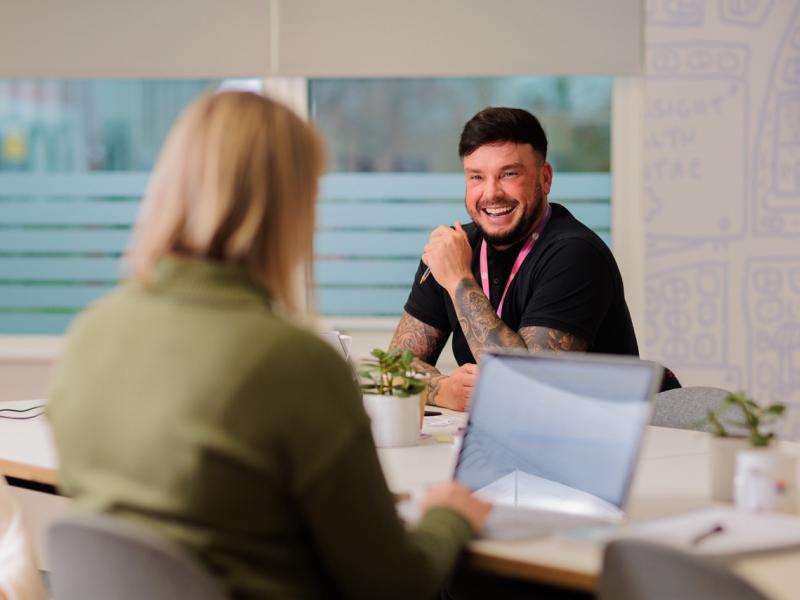 Two people sat at a high desk in an office environment working. The male in the photo is smiling in the direction of the female in a green jumper.
