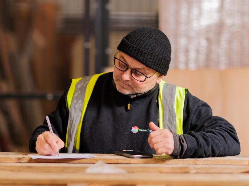 A colleague of One Manchester making notes on top of a stack of building materials. He's wearing a black hat and a high visibility jacket.