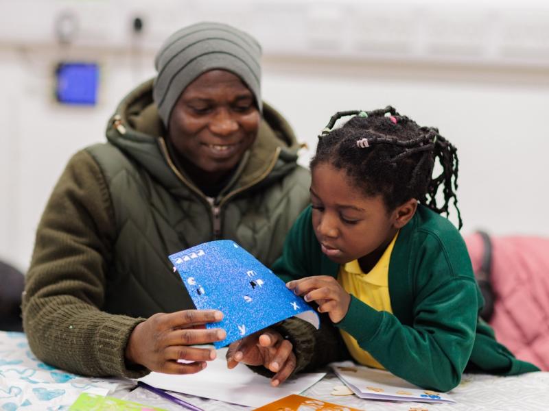 Two people sat doing crafts, one is wearing a green jumper and the other is wearing a jacket and a beanie hat