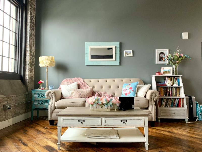 A living room setting which has been recently decorated. The wall is green and the floor in engineered wooden flooring. There is a coffee table in the middle of the room, with a large window to the left allowing light into the room. 