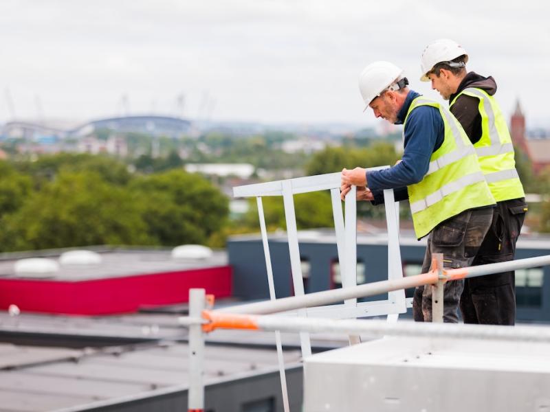 Two One Manchester Colleagues working on a development attempting to fix a post into the top of the building. Both men are wearing high visibility jackets and helmets. The Etihad stadium is in the background of the image.