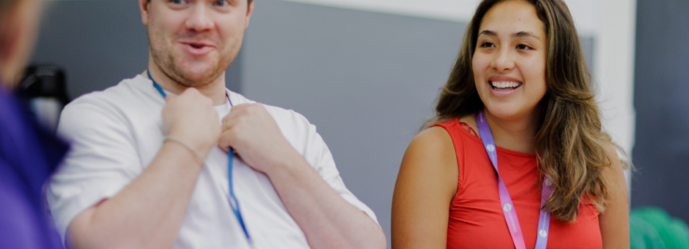 Two people. One is wearing a red dress and the other a white t-shirt sat at table having discussion. 