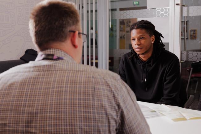 Two people in conversation in an office setting. One is wearing a shirt and the other is wearing a black hoodie. 