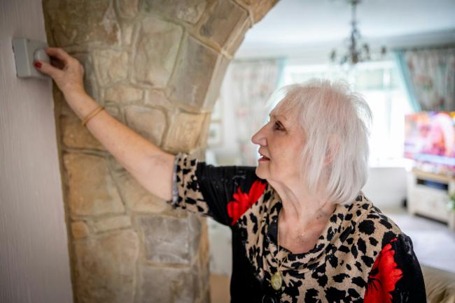 A woman changing the temperature on a thermostat. The woman has grey hair and is wearing leopard print. There is a TV on in the background of the image. 