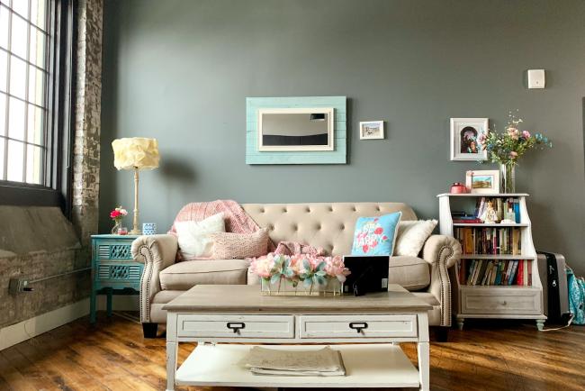 A living room setting which has been recently decorated. The wall is green and the floor in engineered wooden flooring. There is a coffee table in the middle of the room, with a large window to the left allowing light into the room. 